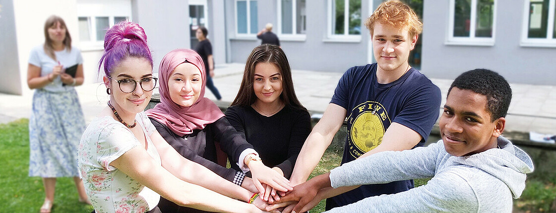 3 Schülerinnen und 2 Schüler stehen im Halbkreis beieinander im Grünen im Hof der Schule und Halten die Hände übereinander. Im Hintergrund steht verschwommen eine Schülerin mit Buch in der Hand. Copyright Caritas Schule Wiener Neustadt